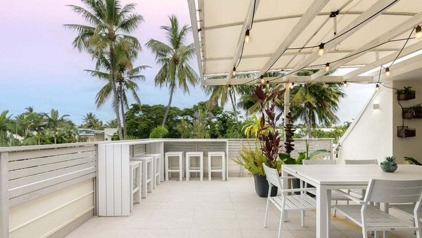 Una terraza en la azotea con una mesa y sillas blancas, rodeada de plantas en macetas y palmeras.
