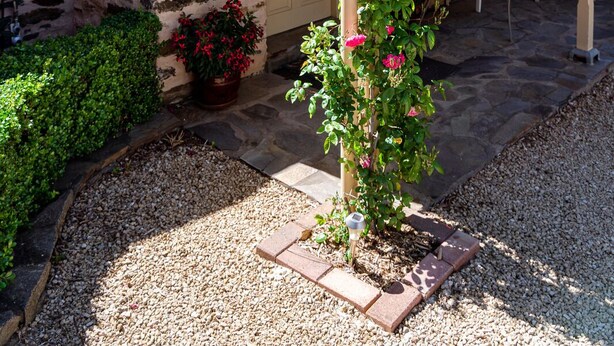 A stone house with a covered patio, a small garden with a rose bush, and a chair and table set up for outdoor dining.