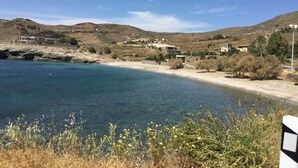 A coastal landscape with a sandy beach, clear blue water, and a hilly backdrop with buildings. - Villa Frear I with infinity pool (KYKLADES)