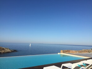 A poolside view with a clear blue sky, a sailboat on the water, and a stone wall. - Villa Frear I with infinity pool (KYKLADES)