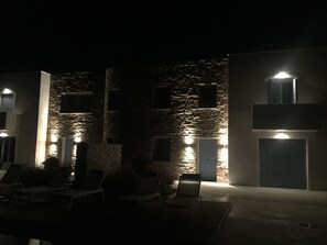 A building with stone walls and multiple windows, some illuminated. There are lounge chairs arranged outside. - Villa Frear I with infinity pool (KYKLADES)