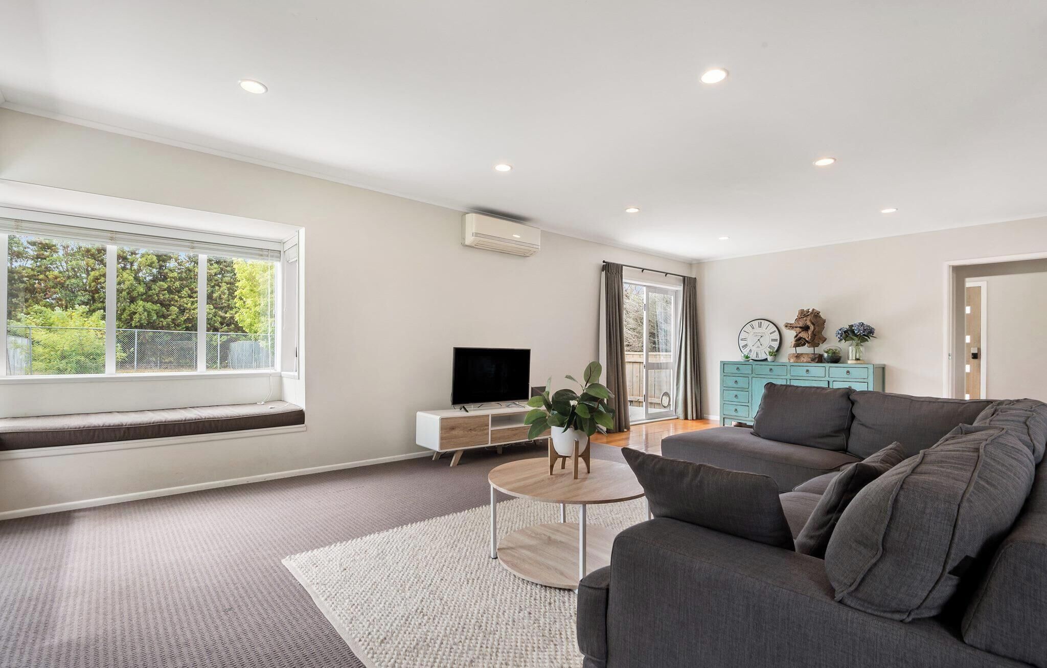 A modern living room with a large window, a flat-screen TV, a grey sofa, a wooden coffee table, and a teal dresser.