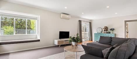 A modern living room with a large window, a flat-screen TV, a grey sofa, a wooden coffee table, and a teal dresser.
