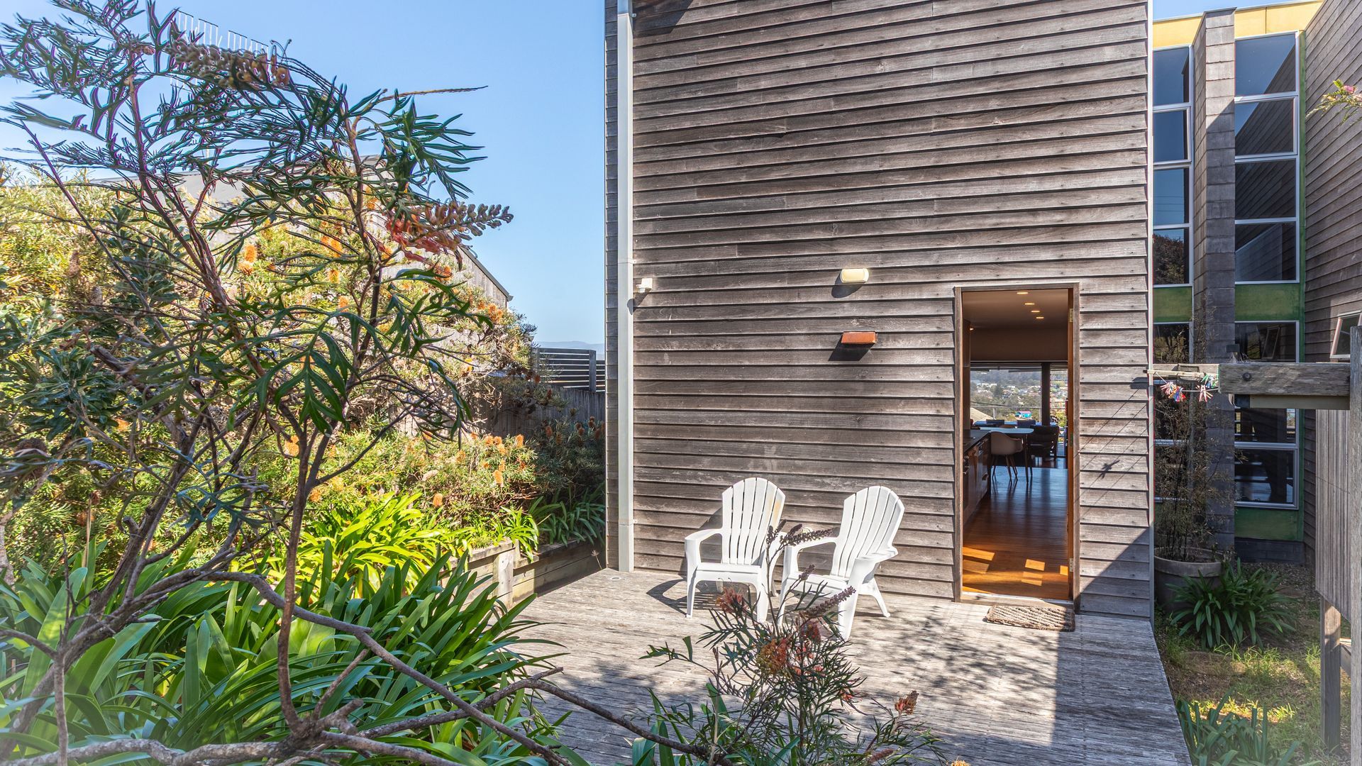 Une maison moderne en bois, dotée d’une terrasse, de deux chaises blanches et offrant une vue sur l’intérieur.