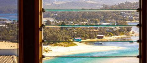 A coastal view through a window with multiple panes, a sandy beach, clear turquoise waters, and a residential area with houses and greenery in the background.