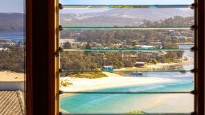 Vista costeira através de uma janela com vários painéis, praia arenosa, águas turquesa cristalinas e área residencial com casas e vegetação ao fundo.