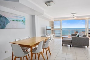 Uma sala de estar moderna com uma mesa de jantar de madeira, cadeiras brancas, um sofá cinzento e uma grande janela com vista para o oceano.