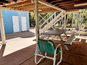 A covered outdoor area with a picnic table and a bench, a blue building with doors, and a staircase leading to an upper level.