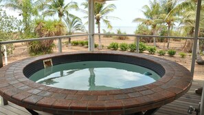 A hot tub with a brick border, surrounded by a wooden deck and metal railing, overlooking a beach with palm trees and the ocean.