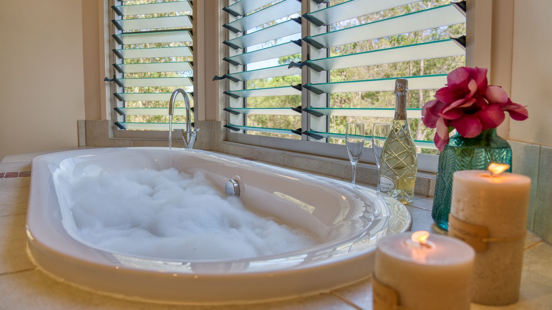 Un baño con una bañera llena de burbujas, una ventana con persianas y velas.
