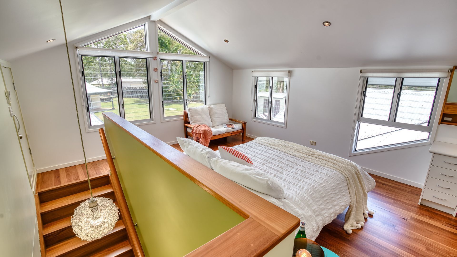 A bedroom with a large bed, a wooden staircase, and multiple windows offering views of the outdoors.