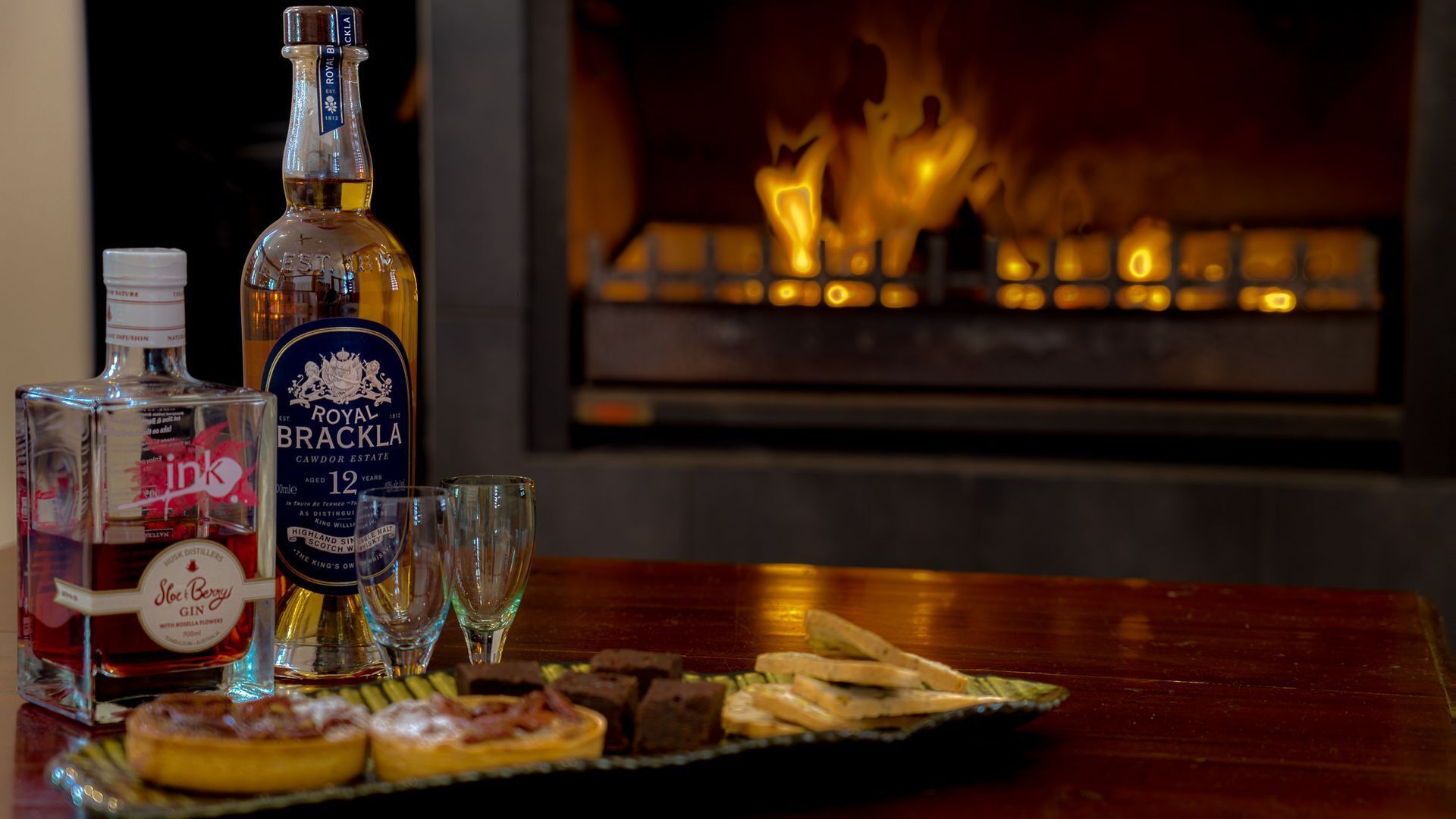 A bottle of Royal Rackla whisky, a bottle of Gin, and glasses on a table with food in front of a fireplace.