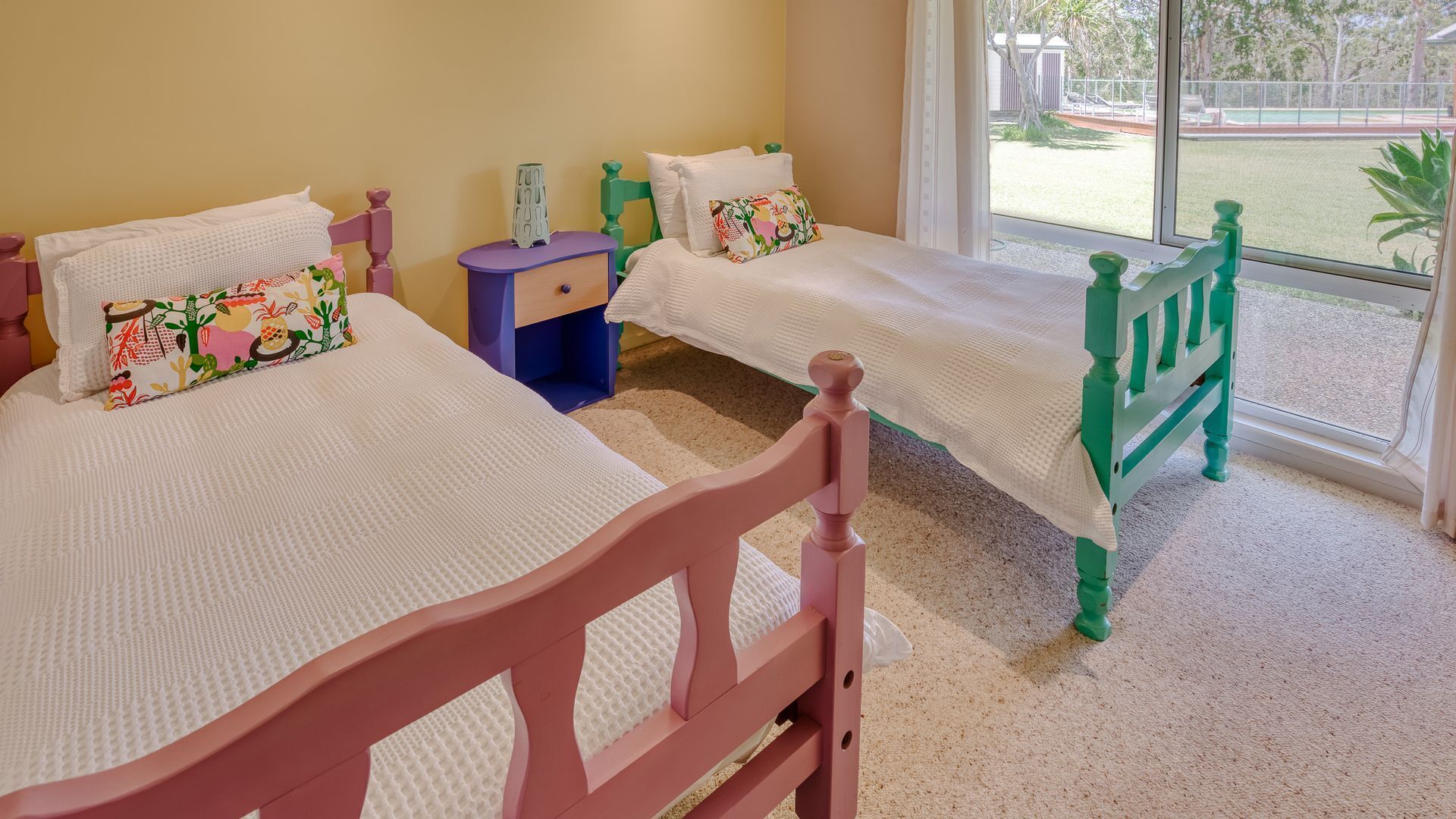 A room with two beds, each with a white mattress and a colorful pillow. There is a small blue nightstand between the beds.