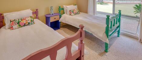 A room with two beds, each with a white mattress and a colorful pillow. There is a small blue nightstand between the beds.