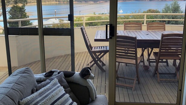 A balcony with a wooden table and chairs, a sofa with cushions, and a view of mountains and a lake.