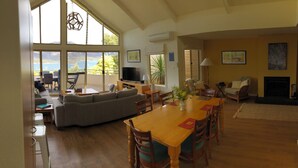 A spacious living area with a large sofa, dining table, and chairs. There is a television, a fireplace, and large windows offering a view of the outdoors.