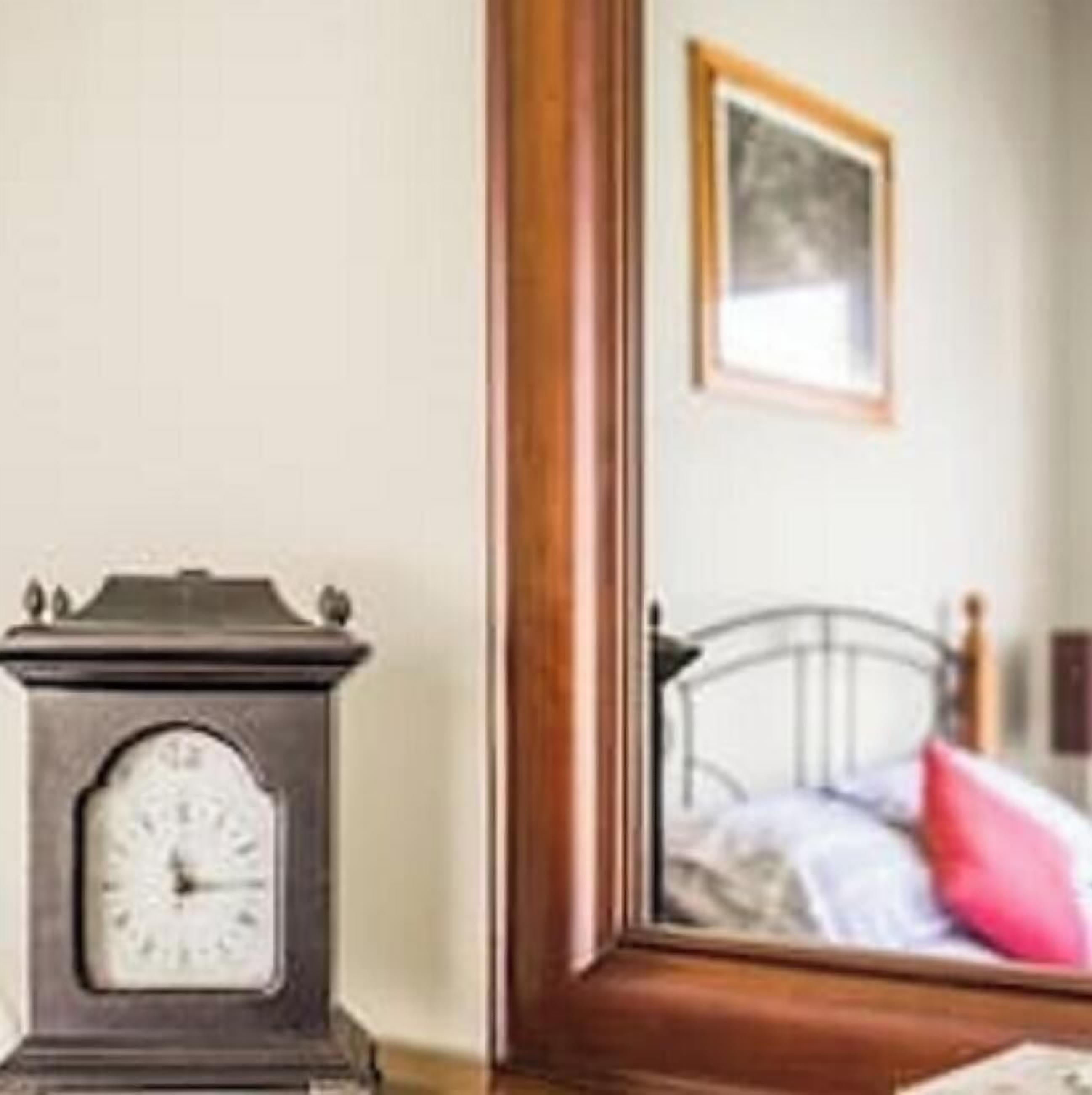 A wooden clock on a table with a mirror reflecting a bed and a picture on the wall.