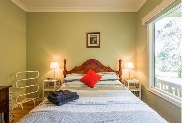 A neatly made bed with a patterned bedspread, two bedside lamps, and a window with a view of trees. - "The Cottage"@ Kilchurn upon Troups (Narre Warren North)