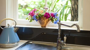 Un évier de cuisine avec un vase de fleurs, une bouilloire et une fenêtre donnant sur un paysage verdoyant.