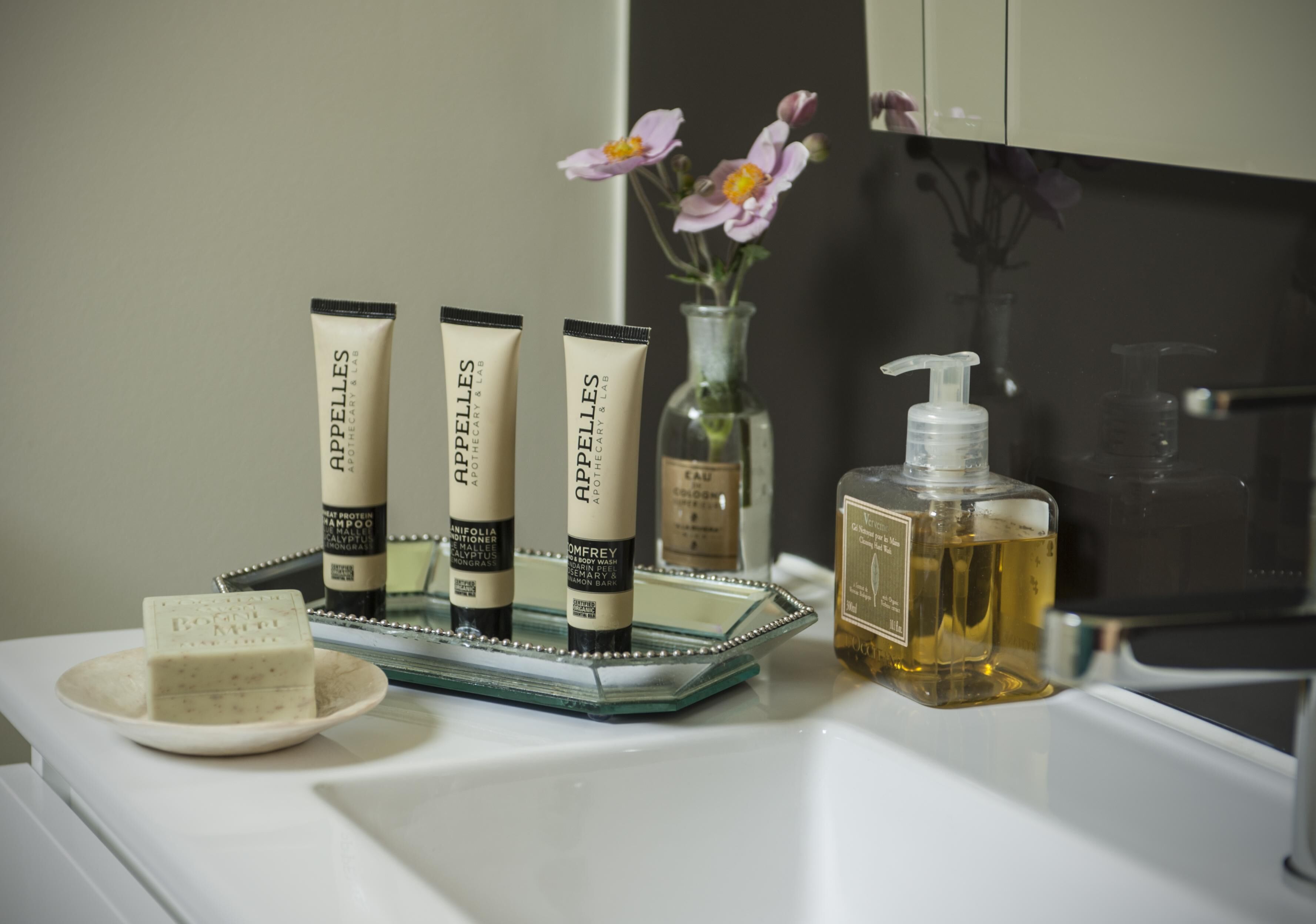 A bathroom vanity with toiletries including a soap bar, three tubes of Aphelis, a glass bottle, and a bottle of liquid soap.
