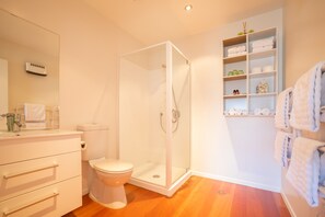 Un baño con inodoro, ducha con mampara de vidrio, lavabo blanco con espejo y repisa con toallas.