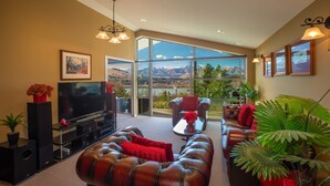 A spacious living room with a large flat-screen TV, leather sofas, and a view of mountains through large windows. - First in the Sun. Quiet 1st floor apartment. Fantastic River and Mountain Views (Frankton)