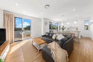 A modern living room with a sofa, coffee table, and a TV. There is a balcony with a view of greenery.