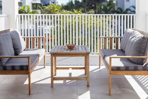 A balcony with a wooden table and two sofas, a white railing, and a view of greenery and buildings.