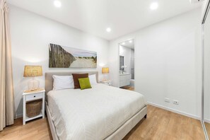 A bedroom with a bed, two bedside tables, a nightstand, a wall-mounted lamp, a painting, and a mirror.