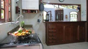 A kitchen with a stove, a fruit bowl, wooden cabinets, a clock, and a window with a view. - Beach house with lots of greenery, a true beach country house.<br> 24-hour security. (Guarujá)