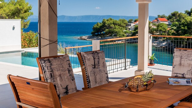Una mesa de madera con un centro de piña, sillas con cojines estampados y vistas a una piscina y al océano.