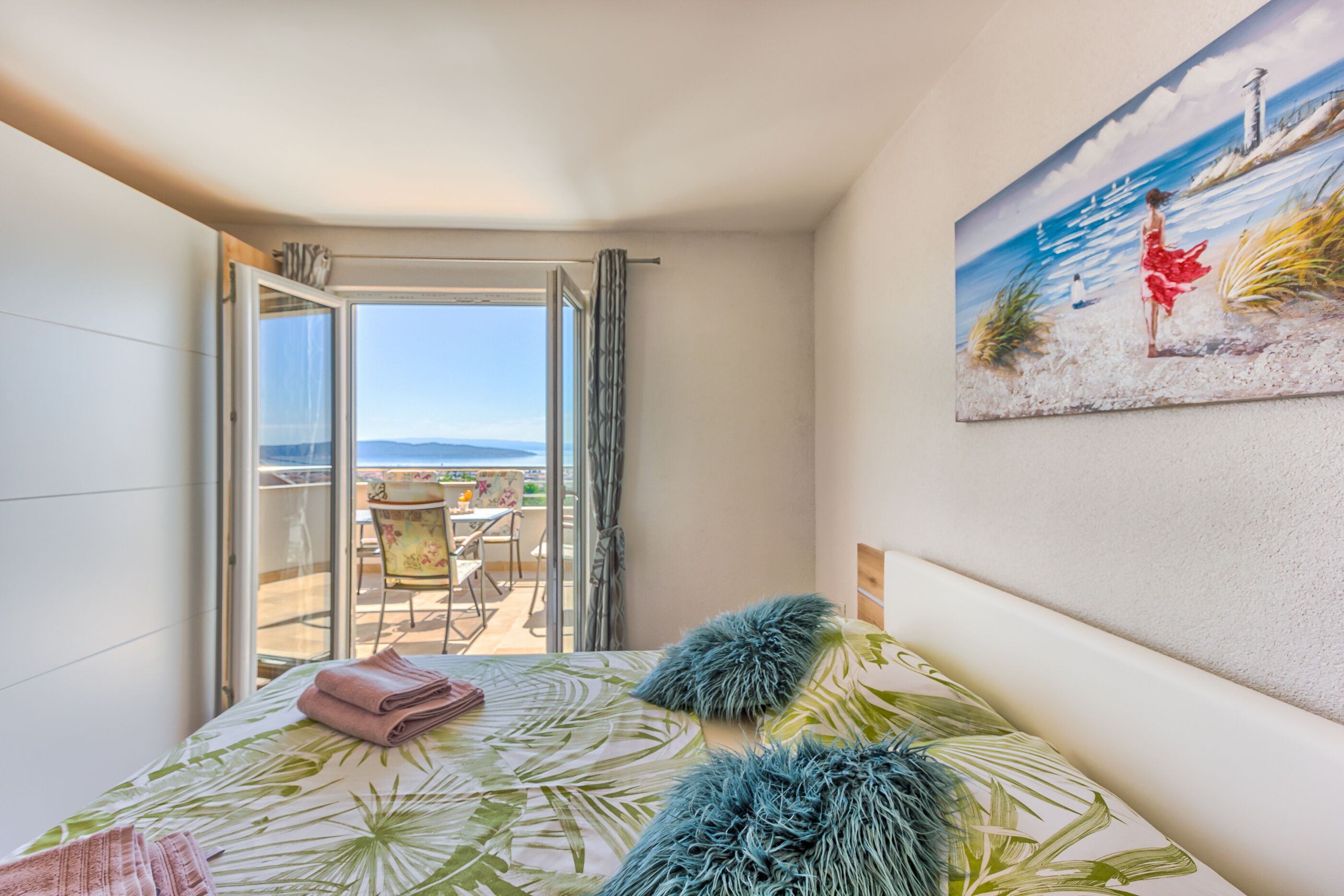 A bedroom with a bed, a painting of a beach scene, and a balcony with a table and chairs.