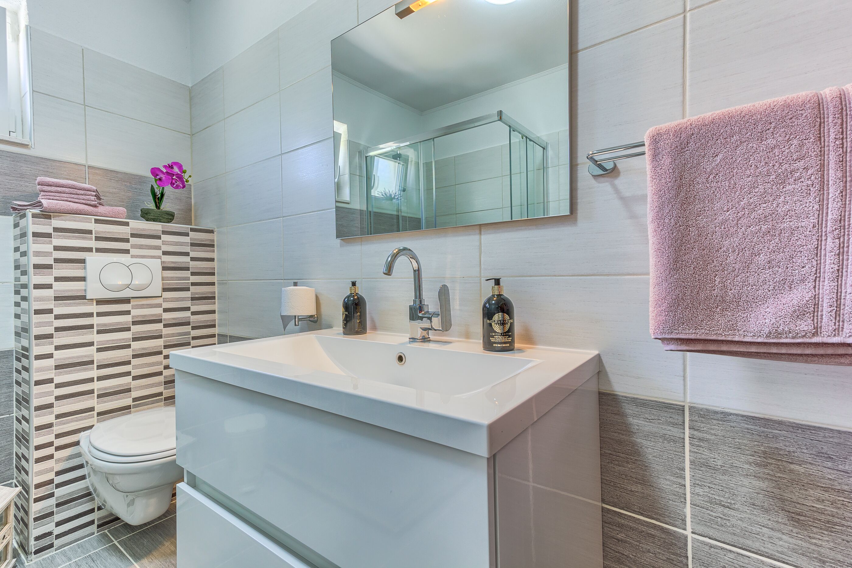 Een moderne badkamer met een witte wastafel, twee kraanwerkens, een spiegel en een roze handdoek op een rek.