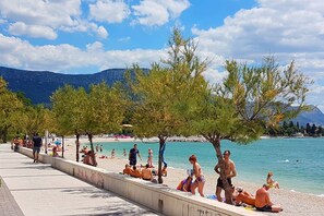 A beachside promenade with trees, people relaxing, and a clear view of the sea and mountains in the background. - Cozy sea view apt. with heated pool and football court (Kaštel Stari)