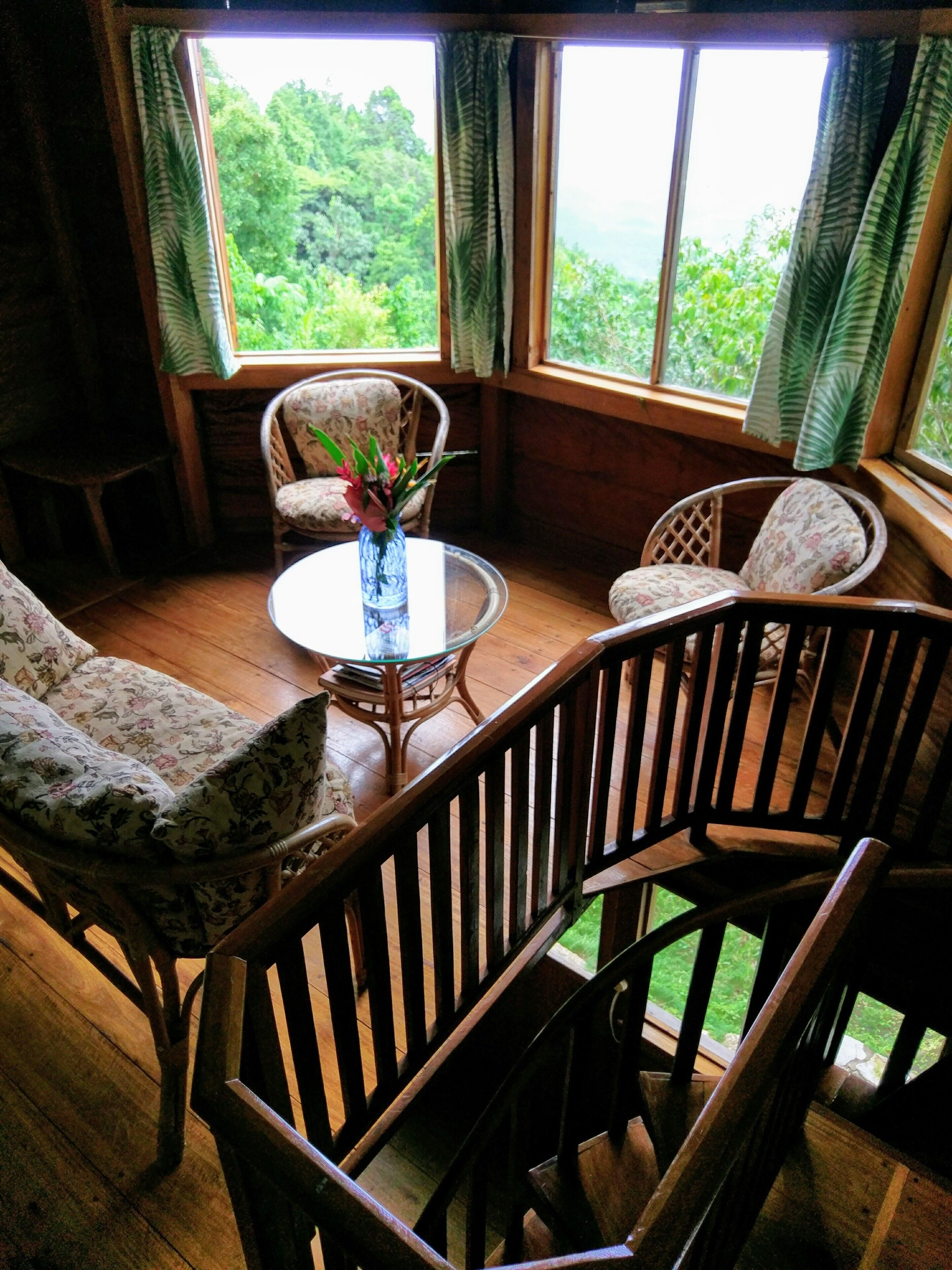 Een gezellige kamer met houten vloeren, een ronde glazen tafel, twee fauteuils met bloemenprint en een bank. Grote ramen met gordijnen bieden uitzicht op groen buiten.
