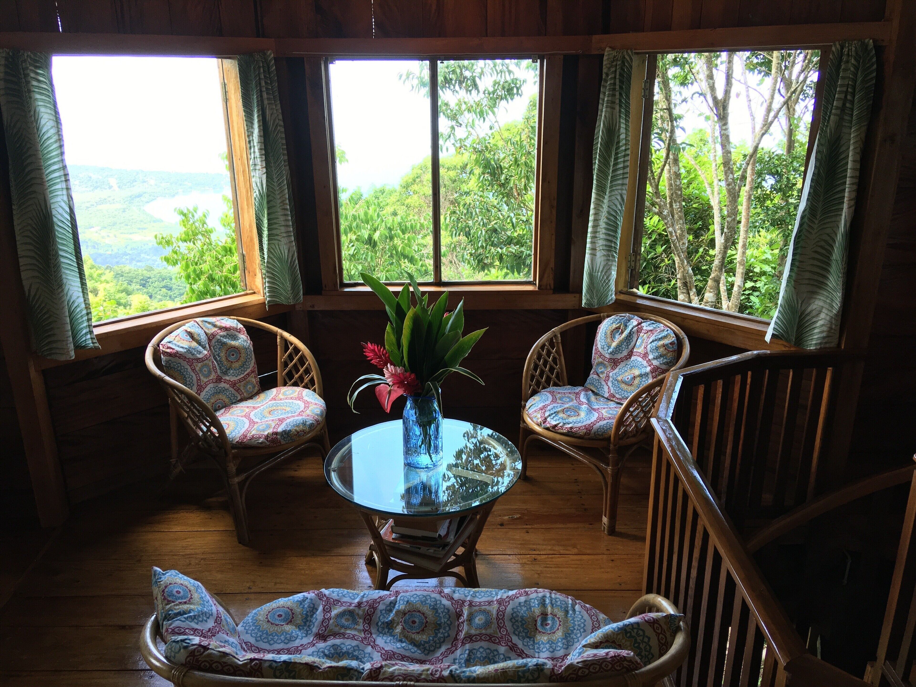 A wooden-floored room with two wicker chairs and a glass table, a vase with flowers, and large windows offering a view of lush greenery.