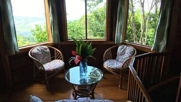 A wooden-floored room with two wicker chairs and a glass table, a vase with flowers, and large windows offering a view of lush greenery.