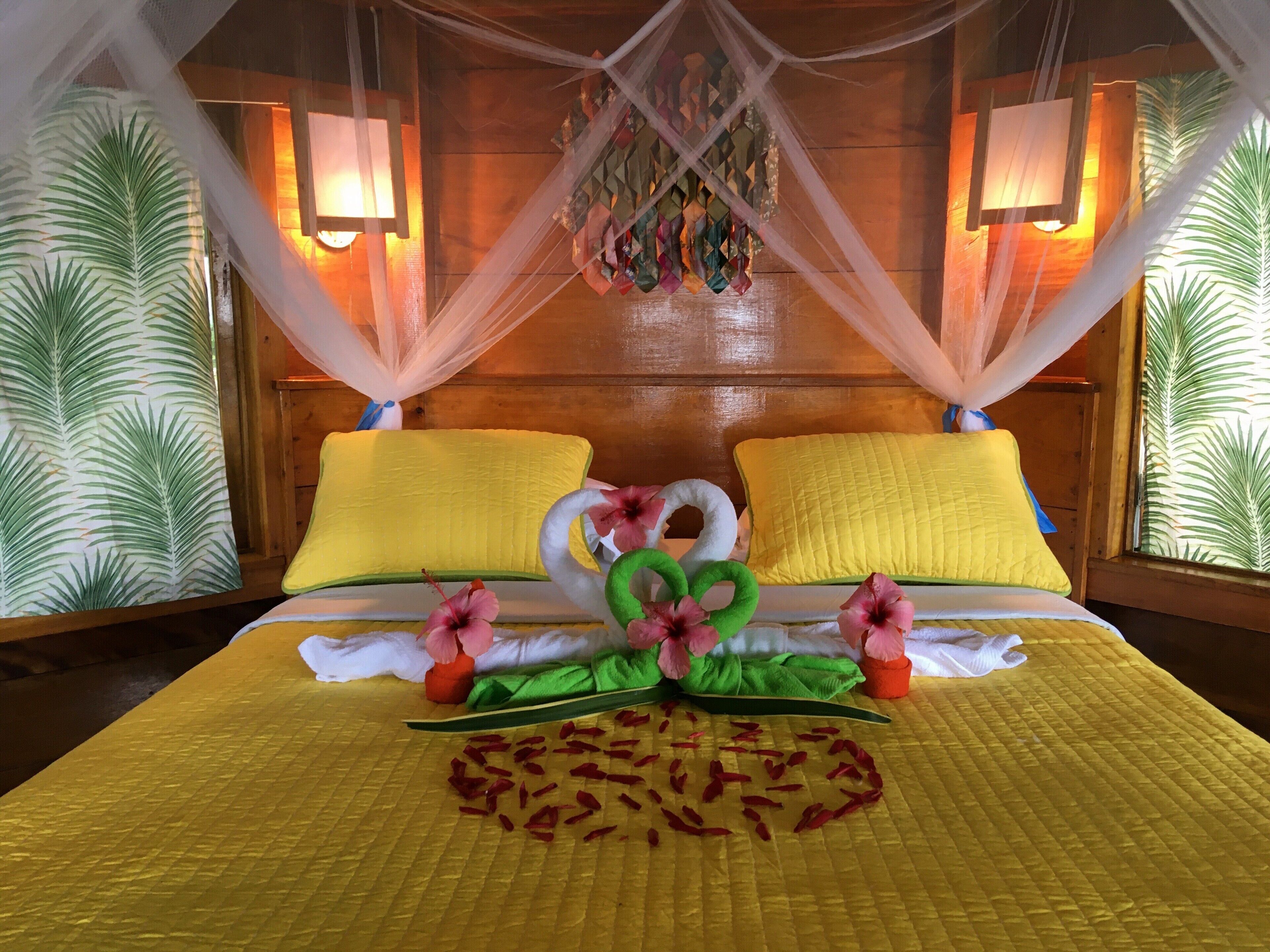 Een hotelkamer met een bed, gele kussens, een baldakijn en een decoratieve bloemen- en bladarrangement.