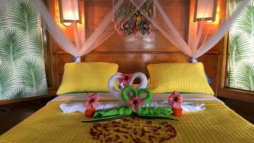 Een hotelkamer met een bed, gele kussens, een baldakijn en een decoratieve bloemen- en bladarrangement.