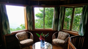 A room with two wicker chairs facing a glass table with a vase of flowers. Large windows with curtains offer a view of greenery outside. - Dragon fly Cottage (Cave)