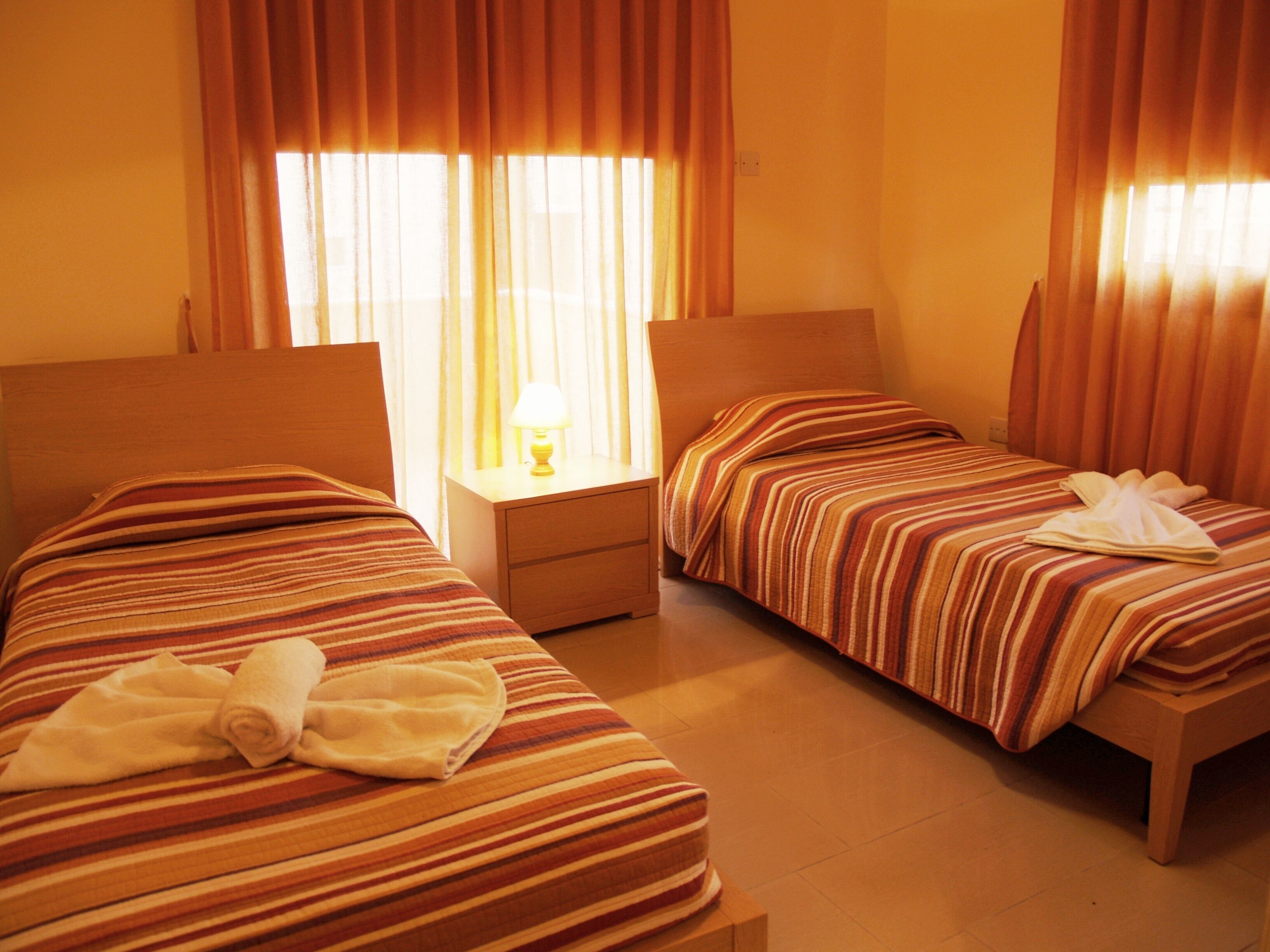 A hotel room with two single beds, a nightstand with a lamp, and a window with curtains.