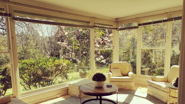 A sunlit room with large windows, a round wooden table with a plant, and a beige sofa. - Lake House Benalla-  Late Sunday checkout available upon request (Benalla)