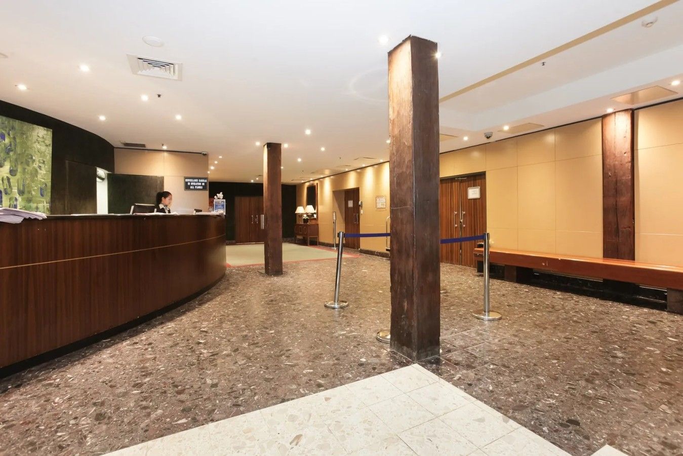 A hotel lobby with a reception desk, marble flooring, wooden pillars, and a seating area.