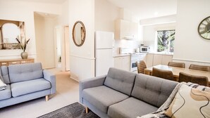 Un salon moderne avec deux canapés gris, une table à manger avec des chaises, un coin cuisine avec des appareils électroménagers blancs et un grand miroir au mur.