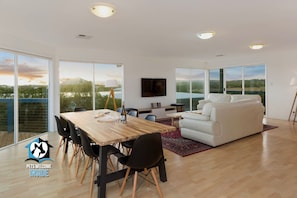 A spacious living area with a wooden dining table, black chairs, a white sofa, and a TV mounted on the wall.
