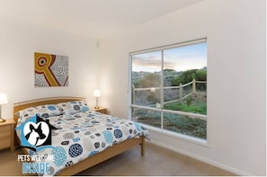 A bedroom with a bed, two bedside lamps, a window with a view of greenery, and a wall art piece.