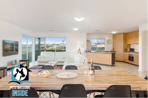 A modern living room with a dining area, kitchen, and large windows offering a view of the city.