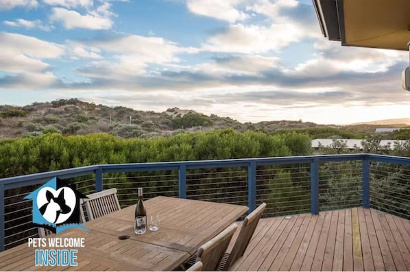 A wooden deck with a table and chairs, overlooking a hilly landscape with dense greenery.