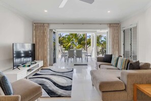 A modern living room with a flat-screen TV, beige sofas, a coffee table, and a view of palm trees through large glass doors.