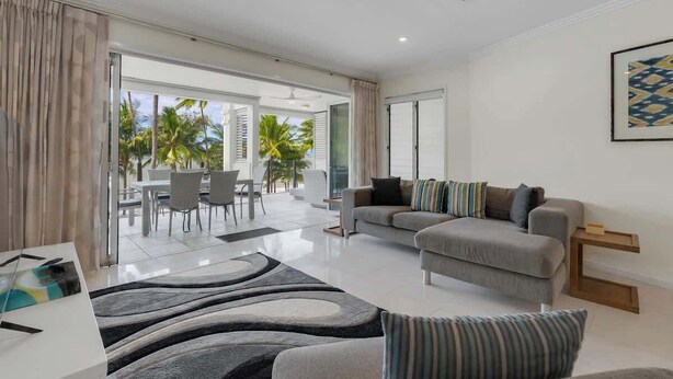A modern living room with a sofa, a dining area with chairs, and a view of palm trees outside. - Mistral on Island Views—Pool Paradise on the Beach (Palm Cove)